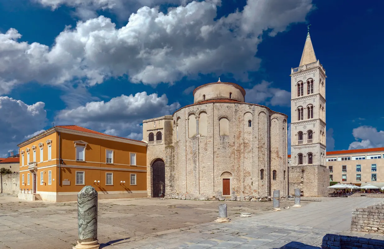 The Church of St. Donatus with its distinctive round shape, standing next to the Roman ruins in Zadar, Croatia. The scene captures the church's ancient stone walls, the adjacent bell tower, and a bright yellow building under a partly cloudy blue sky.