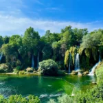 The Kravica Waterfalls in Bosnia and Herzegovina. Lush greenery surrounds the cascading falls, which drop into a clear pool where people can be seen swimming. It is a clear blue sky above and the waterfalls are surrounded by a vibrant landscape, adding to the natural beauty of the location.