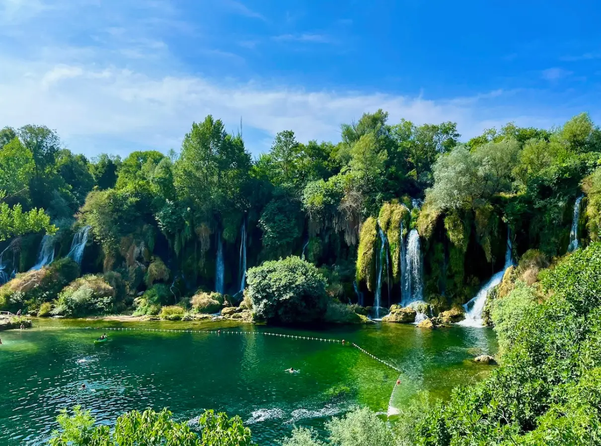 The Kravica Waterfalls in Bosnia and Herzegovina. Lush greenery surrounds the cascading falls, which drop into a clear pool where people can be seen swimming. It is a clear blue sky above and the waterfalls are surrounded by a vibrant landscape, adding to the natural beauty of the location.