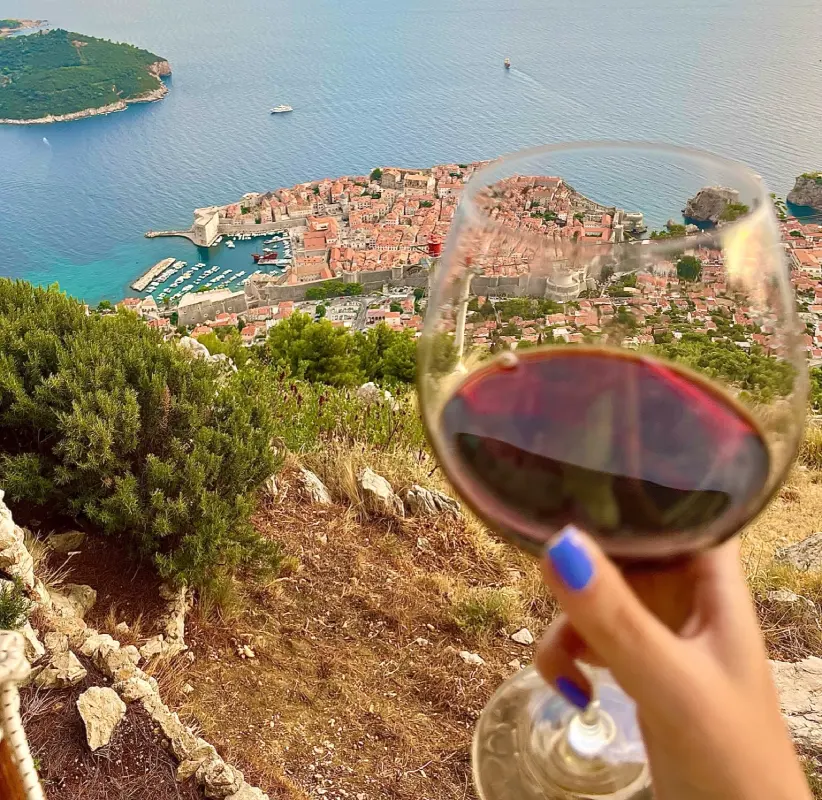 A person with blue-painted nails holds a glass of red wine in the foreground, overlooking a stunning aerial view of Dubrovnik's historic Old Town and the Adriatic Sea in Croatia. Dubrovnik's terracotta rooftops can be seen in the background. 