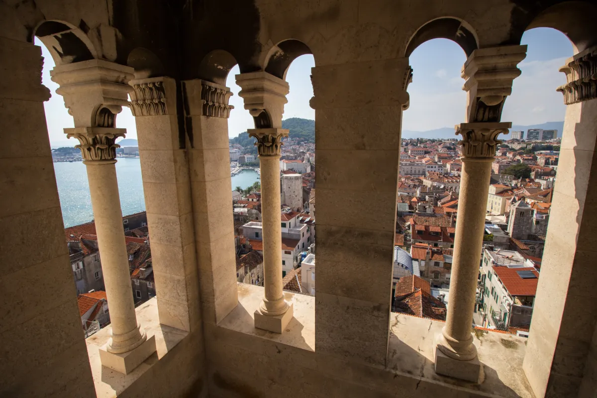 The view from the bell tower of Saint Domnius Cathedral in Split, Croatia. It showcases thr ornate stone columns framing a panoramic view of the city’s terracotta rooftops and the Adriatic Sea in the distance.