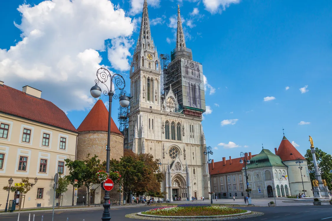 The Cathedral of Zagreb in Croatia that showcases its twin spires, one of which is under renovation and covered with scaffolding. The background scene includes surrounding buildings, a flower bed, and a bright blue sky with scattered clouds.