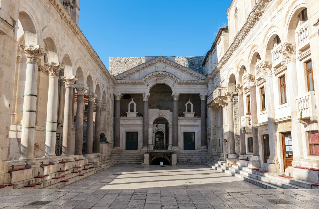 The Peristyle of Diocletian's Palace in Split, Croatia, a Roman structure featuring a grand courtyard surrounded by columns and arched entrances, leading to an ornate facade with twin bell towers. A clear blue sky can be seen above