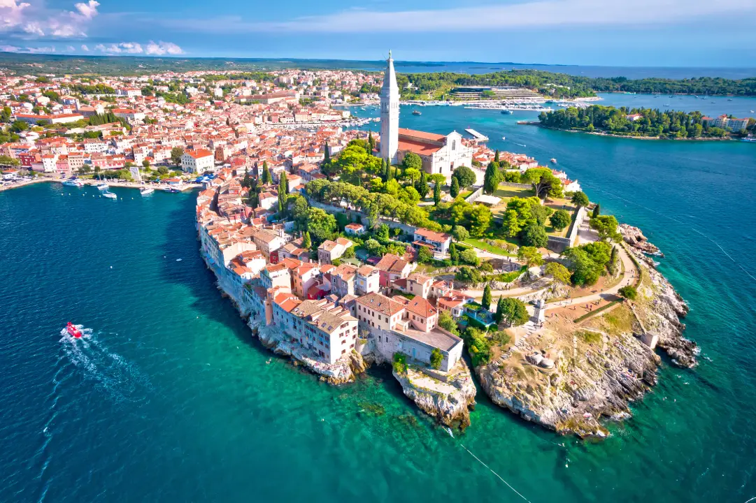 Aerial view of Rovinj Old Town, featuring a dominant church with a tall bell tower surrounded by lush greenery, historic buildings, and the Adriatic Sea. The terracotta rooftops are also on display next to the surrounding crystal-clear blue waters.