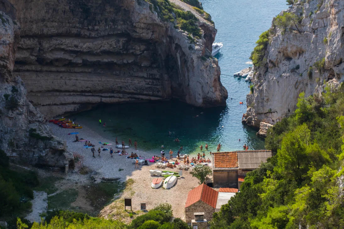 Aerial view of Stiniva Cove on Vis Island, Croatia, showcasing a secluded pebble beach surrounded by high cliffs with a clear turquoise sea. People are sunbathing, swimming, and enjoying water activities, with kayaks and paddleboards visible near the shore.