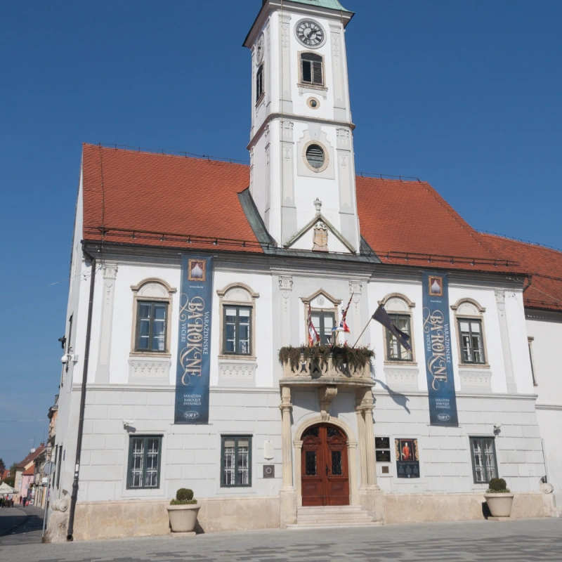 The exterior of Varaždin Town Hall features a white facade and a clock tower adorned with banners on the building that is advertising for an event. The building features ornate architectural details, a red-tiled roof, and a central balcony with flags. 