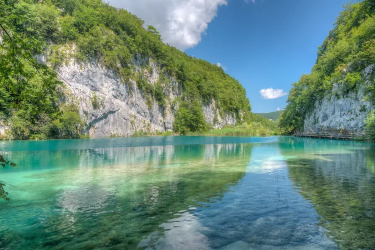 Clear turquoise water reflects lush green hills and rocky cliffs under a bright blue sky with a few clouds, capturing a beautiful Croatian landscape.