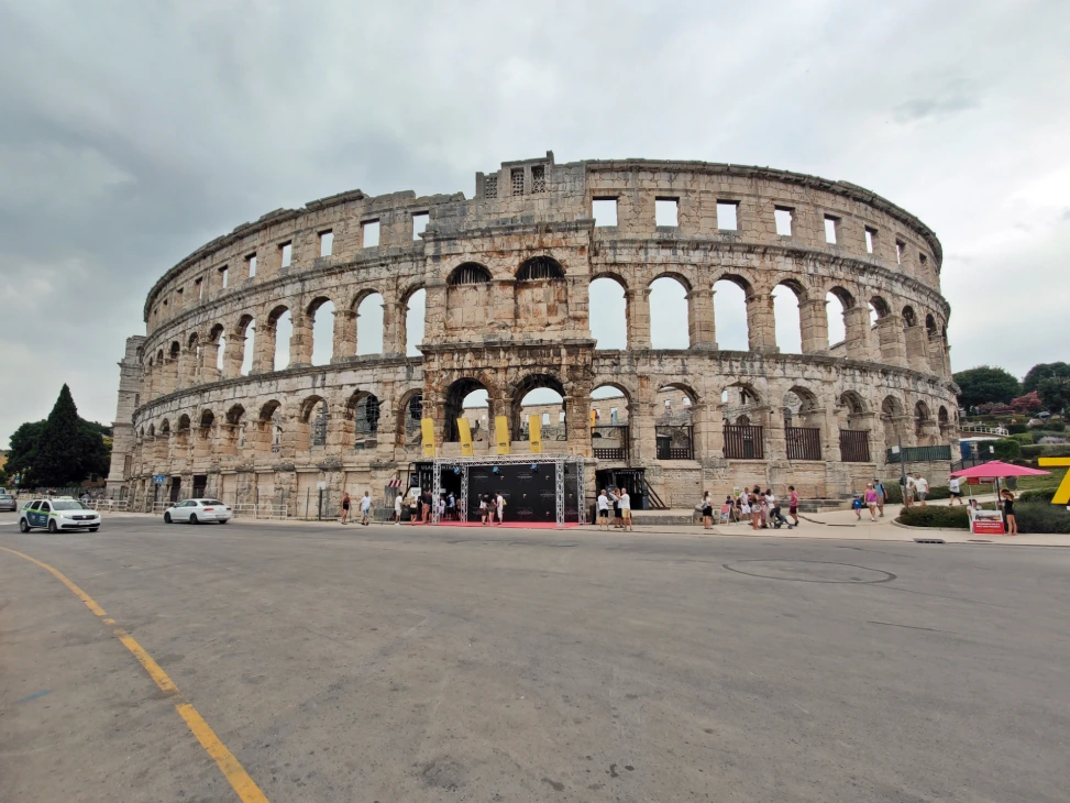 Outside view of the Pula Arena in Croatia on a cloudy day, showcasing its ancient Roman architecture with multiple arched openings and weathered stone walls. People and cars can be seen in front.