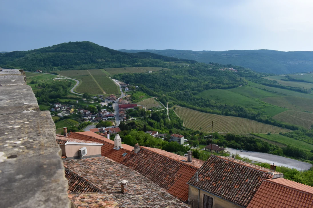 A panoramic view under a partly cloudy sky from the walls of Motovun Castle, overlooking the Mirna River valley with rolling hills and vineyards. The foreground features the terracotta rooftops of nearby houses while winding roads and clusters of buildings are scattered across the landscape. 