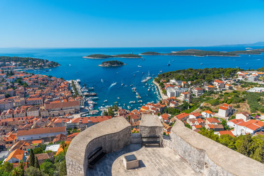 A panoramic view from the Fortica Fortress in Hvar, Croatia. The image captures the blue waters of the Adriatic Sea dotted with small boats, surrounded by lush green islands. Below, the town of Hvar is visible, with its terracotta rooftops and narrow streets. The Paklinski Islands can be seen from a far distance. 