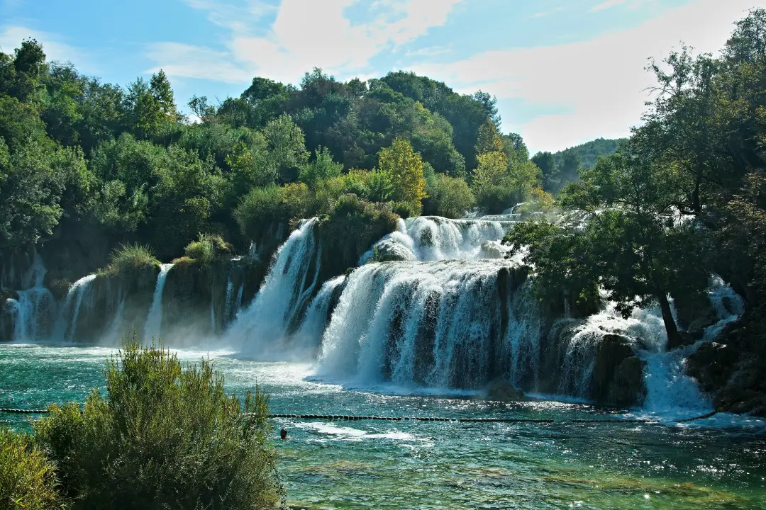 A scenic view of the Roski Slap waterfalls in Krka National Park, Croatia, with multiple cascades surrounded by lush green trees under a bright sky. A person can be seen swimming in the clear turquoise waters.