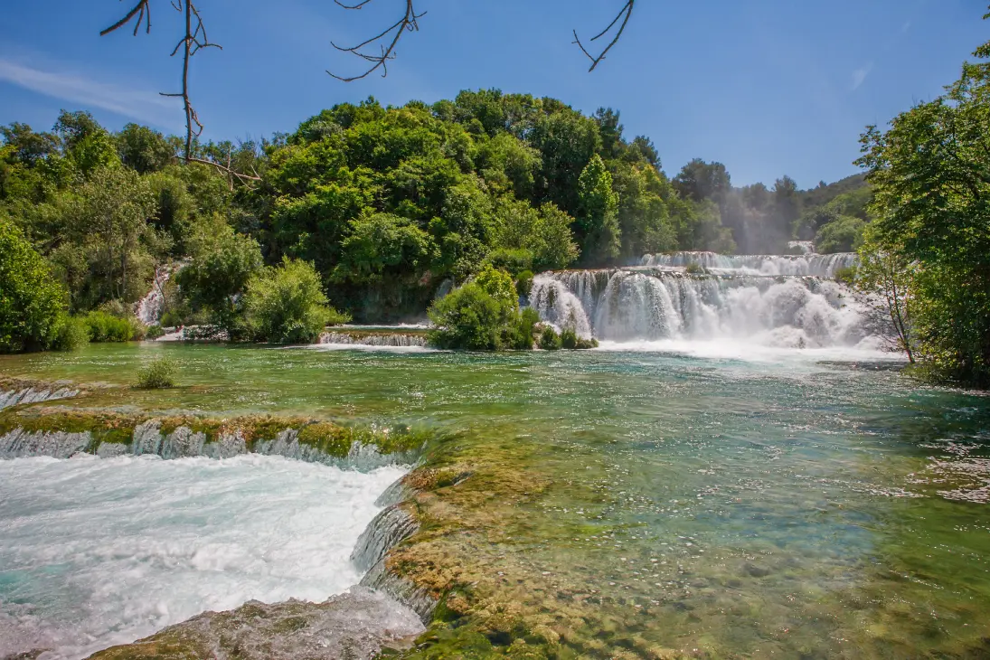 A scenic view of Skradinski Buk, a waterfall in Krka National Park, Croatia, with clear turquoise water cascading over moss-covered rocks, surrounded by lush green trees under a bright blue sky.