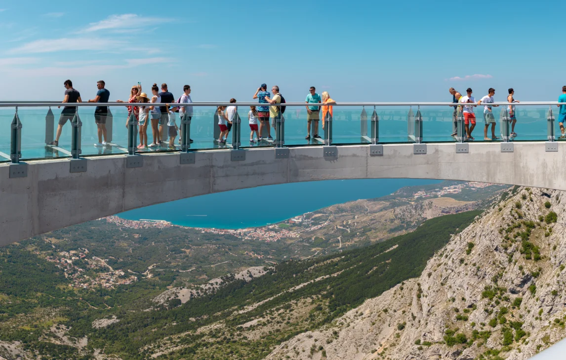 A group of tourists walking on the Skywalk Biokovo, a glass walkway extending over a steep cliff, with a stunning view of the Adriatic Sea and the surrounding mountainous landscape of Biokovo National Park in the background.