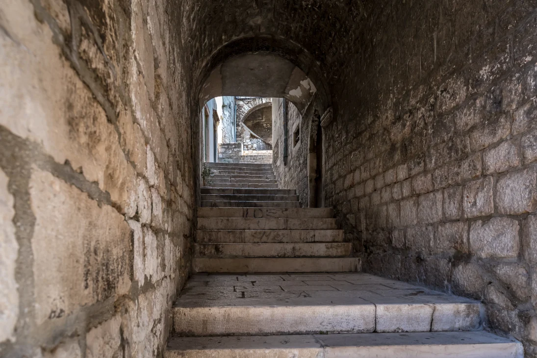 Stone stairs lead up through a narrow, arched passageway in Šibenik, Croatia. To the side you can see aged, textured walls with sunlight illuminating the path ahead. 