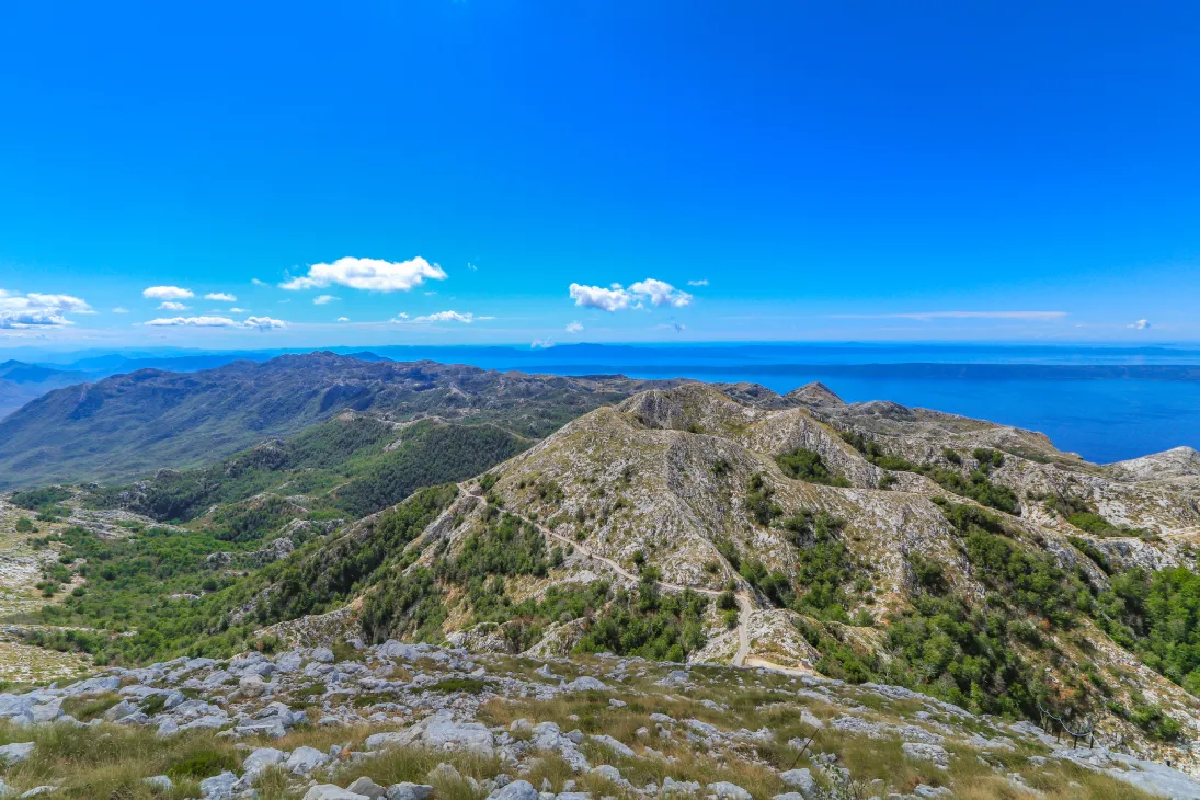 A view from the Sveti Jure peak in Biokovo Nature Park showcasing rocky mountain terrain and lush green patches, with the Adriatic Sea and clear blue sky in the background.