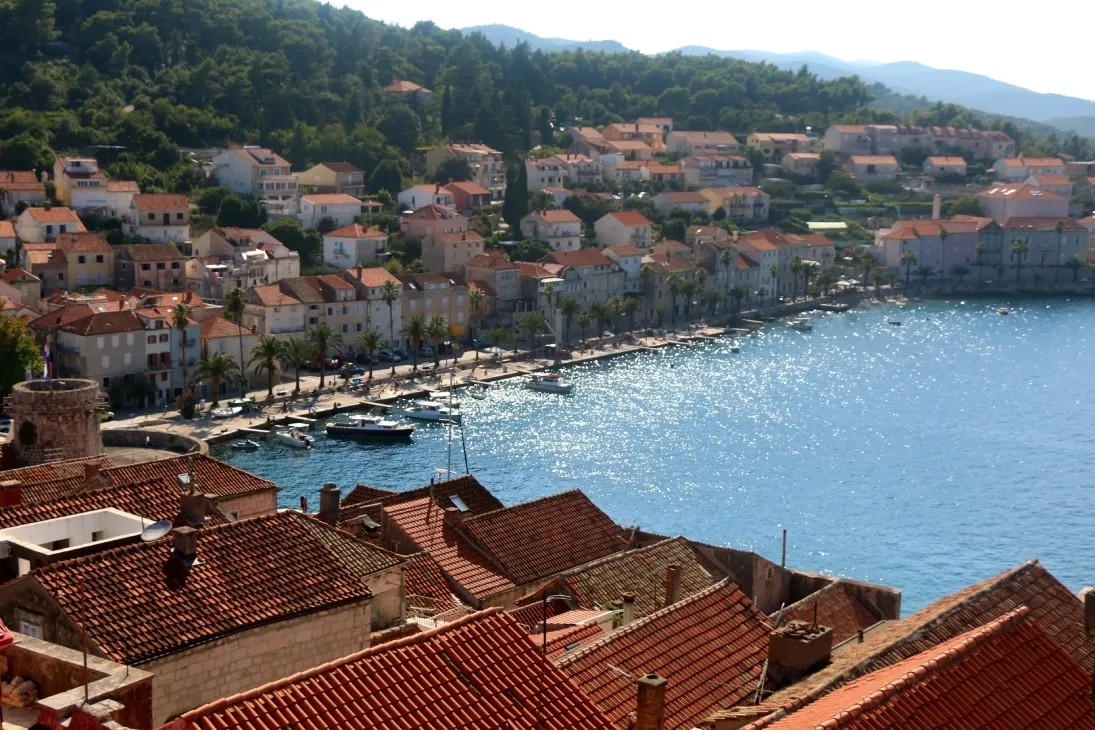 View from St. Mark's Cathedral Bell Tower in Korčula, showcasing terracotta rooftops and a scenic coastal town with clear blue waters and boats docked along the shoreline.