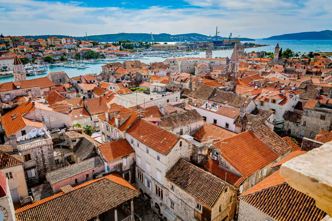 A panoramic view of Trogir, Croatia, with a dense cluster of buildings featuring red-tiled roofs from the Cathedral of St. Lawrence. The scene includes a backdrop of a harbour filled with boats and the blue sea extending to the horizon, surrounded by hilly terrain. 