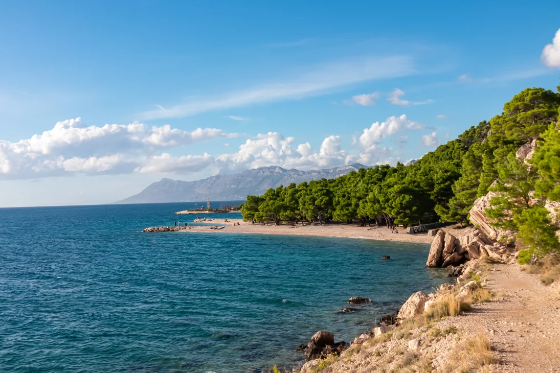 A beautiful view of Ramova Beach in Makarska, Croatia, showcasing the clear blue waters of the Adriatic Sea, a rocky shoreline, and lush green pine trees under a partly cloudy sky with distant mountains in the background.