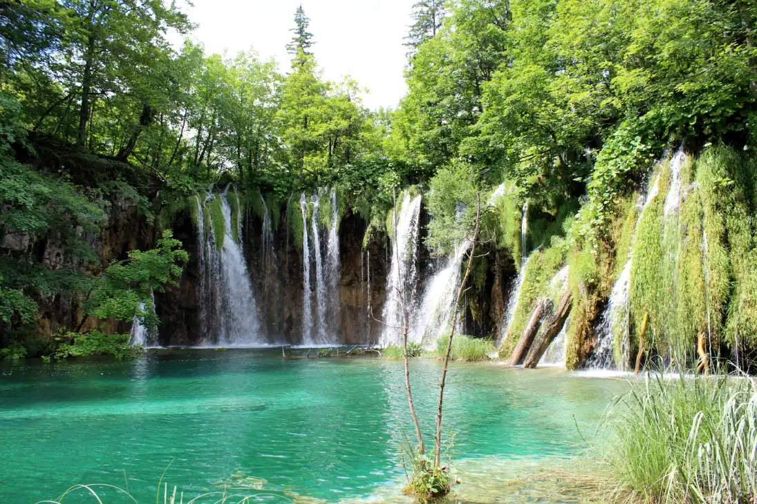 A series of cascading waterfalls flow into a turquoise lake, surrounded by lush green foliage in Plitvice Lakes National Park, Croatia.