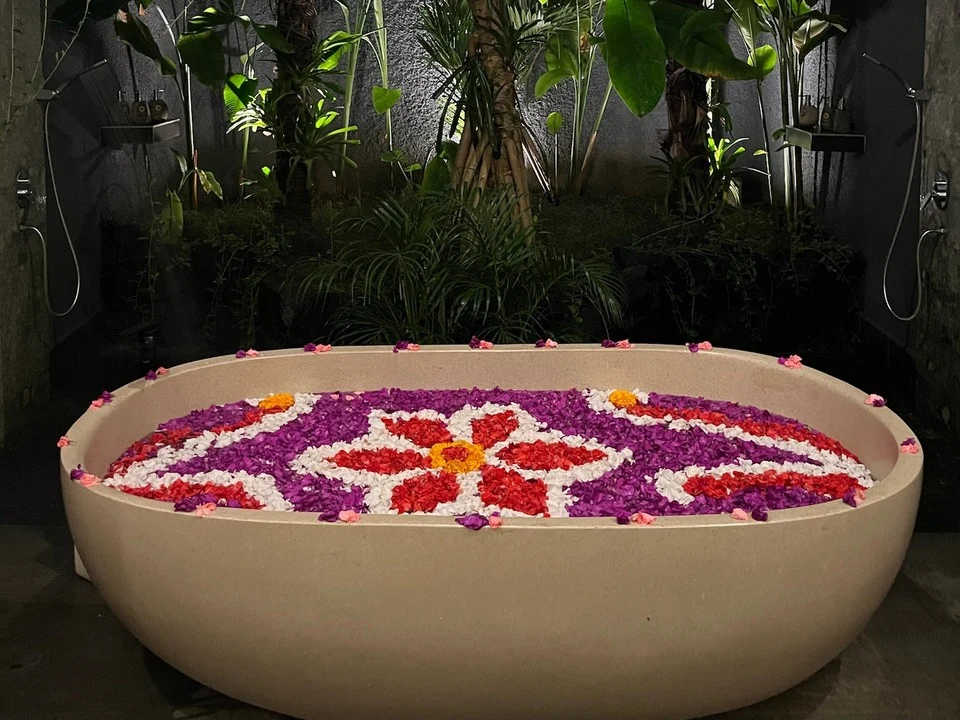 An outdoor stone bathtub filled with colourful flower petals arranged in a symmetrical floral pattern, including vibrant red, purple, white, and orange hues. The bath is at nighttime in a tropical environment, surrounded by lush green plants and softly lit for a tranquil ambience. 