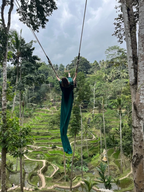 A woman in a flowing green dress swings high above the lush, rice terraces of Tegallalang in Bali. 