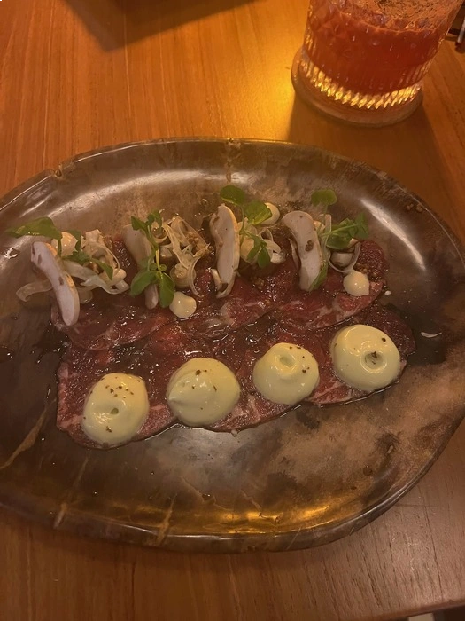 A plated dish featuring thinly sliced beef carpaccio, topped with microgreens, mushrooms, and small dollops of creamy garlic sauce. The dish is served on a rustic dark plate, with a cocktail glass containing a vibrant orange drink visible in the background.