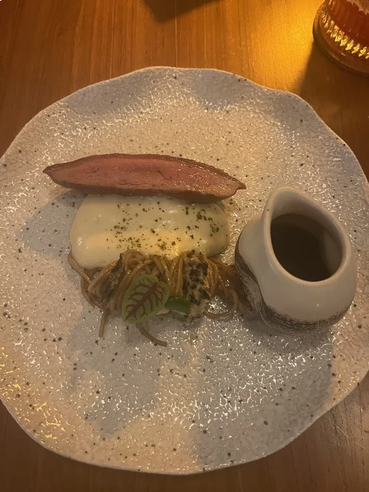 A plated dish featuring a slice of seared duck placed atop a creamy sauce and noodles garnished with microgreens. To the side, there is a small ceramic pitcher, containing a sauce. The dish is served on a textured, irregularly shaped plate.