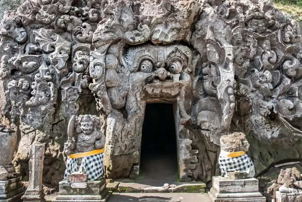 The stone entrance of Goa Gajah is carved with a large, fierce face featuring bulging eyes and an open mouth that serves as the doorway. 