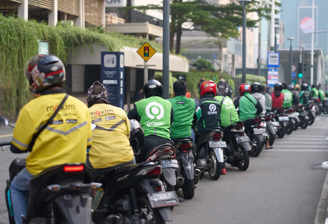 A long line of motorbike drivers, mostly wearing green Gojek jackets and helmets, waits on the street in Indonesia. 