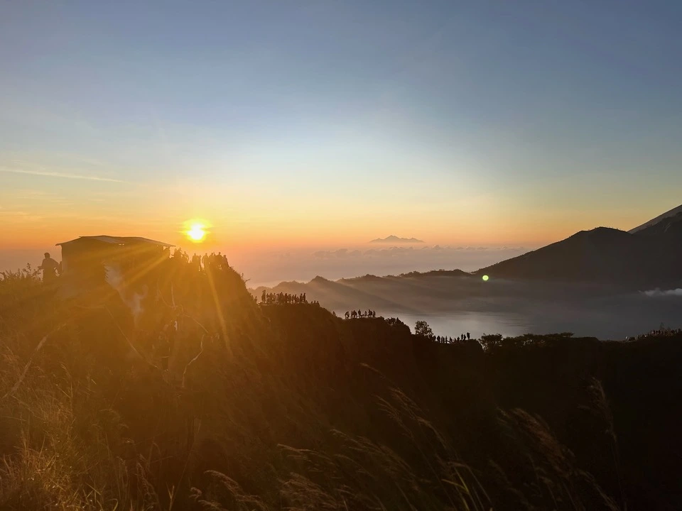 A stunning sunrise view at Mount Batur in Bali, Indonesia. The golden sun rises above a misty horizon next to a crowd of people gathered at the ridge to enjoy the breathtaking scenery.