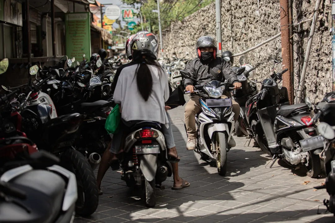 A narrow, crowded alleyway in Bali, Indonesia, where two people on scooters face each other next to tightly parked motorbikes lining both sides. The riders, wearing helmets, carefully navigate the confined space, with one person dressed in a black jacket and the other in a white shirt holding a green bag