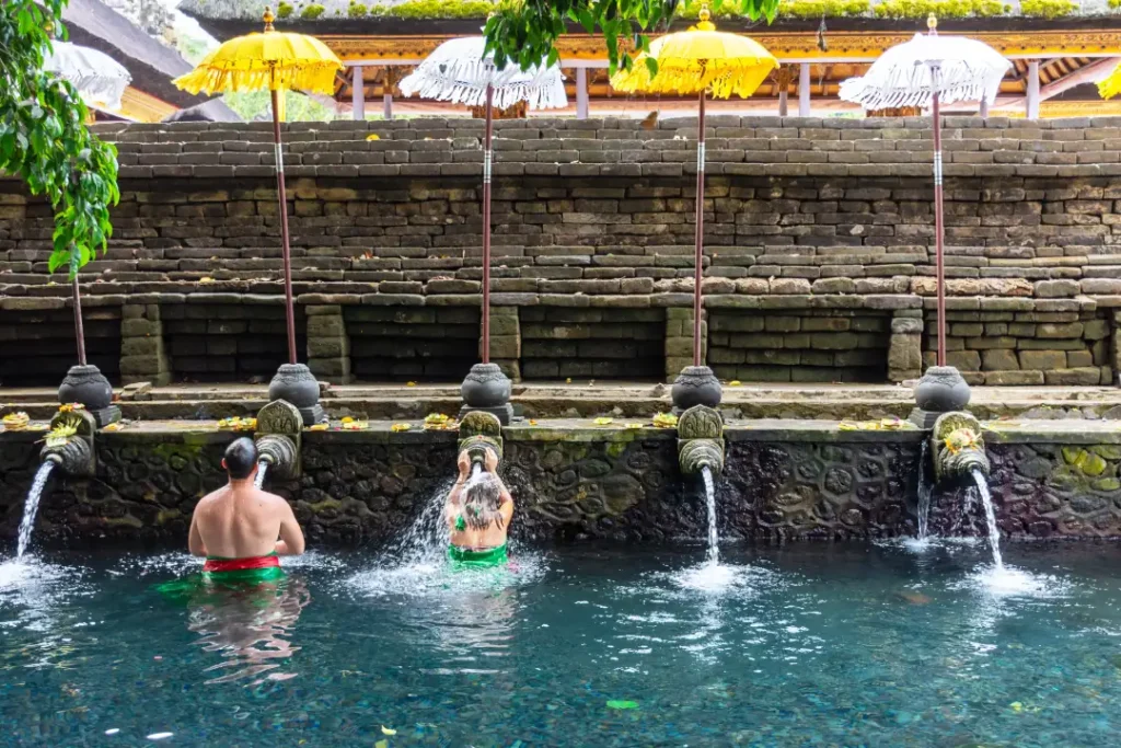 Two people engaging in a purification ritual at a traditional Balinese water temple, standing in a clear pool under ornate stone spouts with water flowing over them.