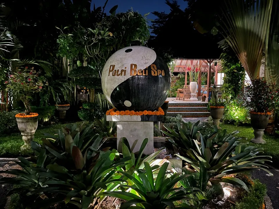  An outdoor setting featuring a stone sculpture with a yin-yang design and the text "Putri Bali Spa" in gold letters. The sculpture is surrounded by lush greenery, potted plants, and vibrant marigold decorations.