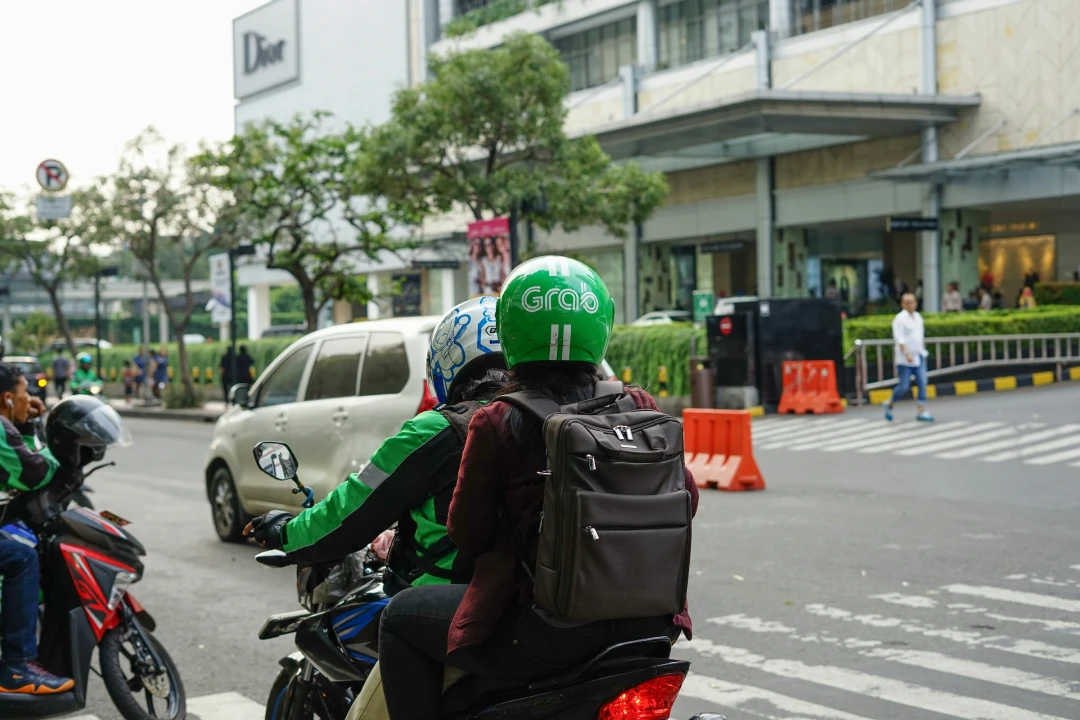 A GrabBike driver wearing a green jacket and helmet with "Grab" branding is giving a ride to a passenger with a backpack. They are navigating a busy street near a shopping area, with vehicles and pedestrians in the background.