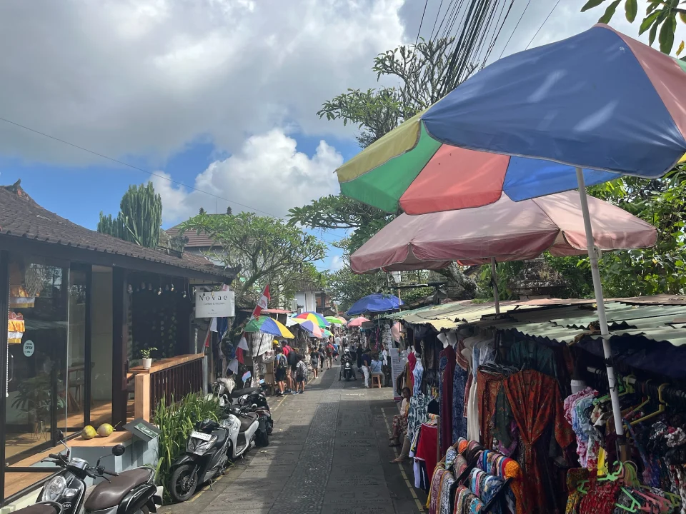 A vibrant street at the Ubud Art Market in Bali, Indonesia. Brightly coloured umbrellas provide shade for market stalls displaying an array of fabrics and handmade goods. Motorbikes are parked along the side, and the scene is framed by traditional Balinese architecture and lush greenery under a partly cloudy sky.