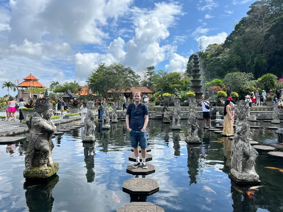 Someone standing on stepping stones surrounded by statues in a reflective pond at Tirta Gangga Water Palace in Bali, Indonesia on a east bali tour. By the side is greenery, traditional Balinese architecture with red-roofed pavilions, and visitors exploring the tranquil water garden. 