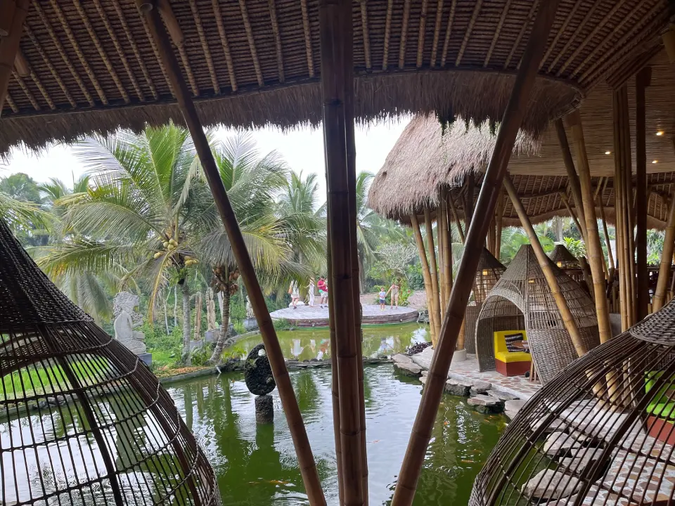 An open-air restaurant with bamboo architecture, overlooking a tropical scene in Bali. The foreground has hanging wicker seats and bamboo columns, while the background has a pond surrounded by palm trees and greenery. 