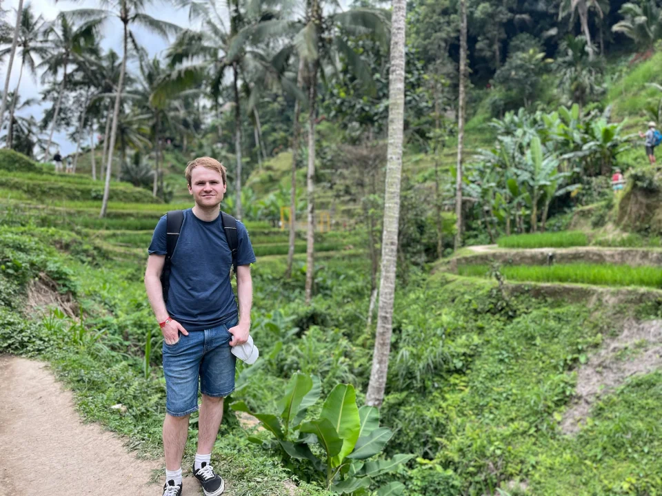 Standing on a walking path through lush, terraced rice fields at the Tegallalang Rice Terraces in Bali. Surrounded by vibrant greenery, tall palm trees, and layered rice paddies.