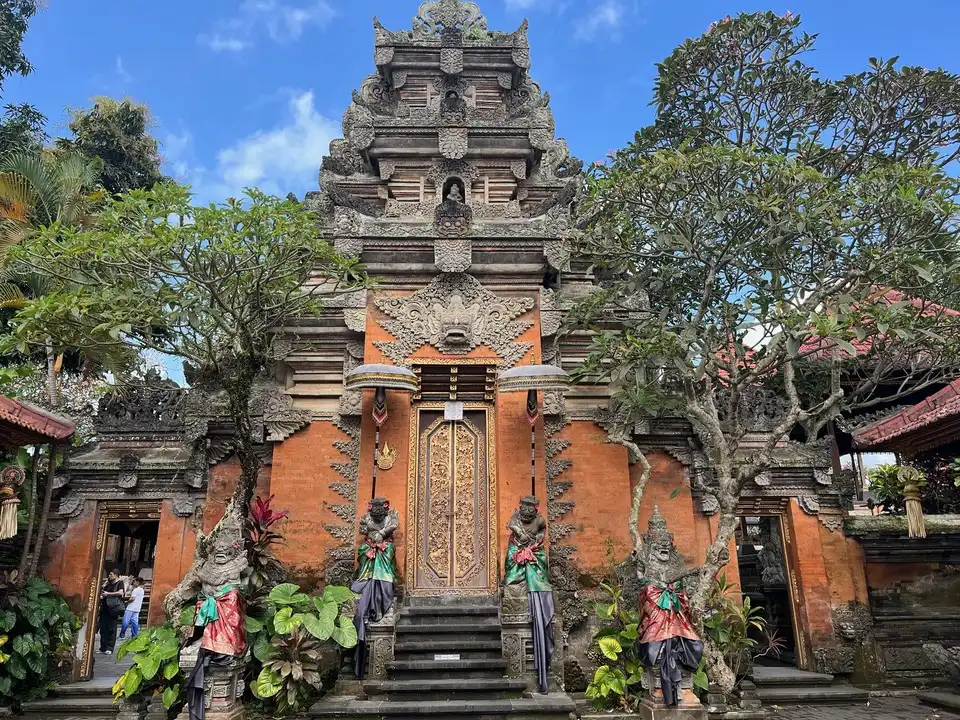 An intricately designed entrance of Ubud Palace (Puri Saren Agung) in Bali, Indonesia.  featuring detailed stone carvings, a golden decorative door, and statues surrounded by greenery and tropical plants.