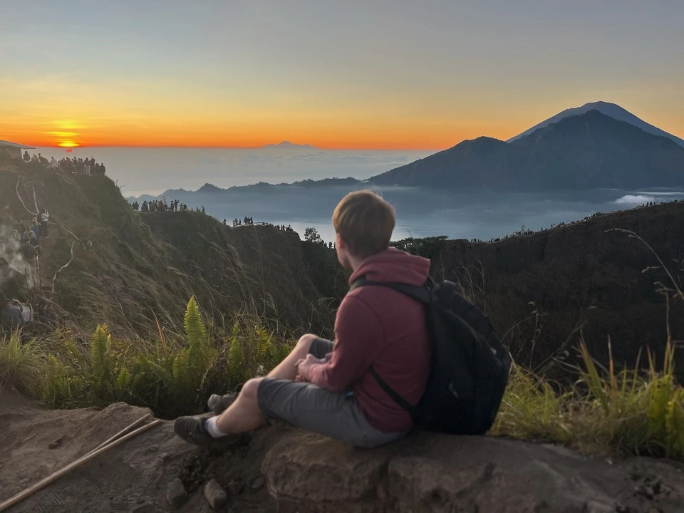 A person sitting on a rocky ledge overlooking a sunrise view from Mount Batur in Bali, Indonesia. The scene includes a vibrant orange and yellow sunrise, distant mountain peaks, and a valley filled with morning mist. 