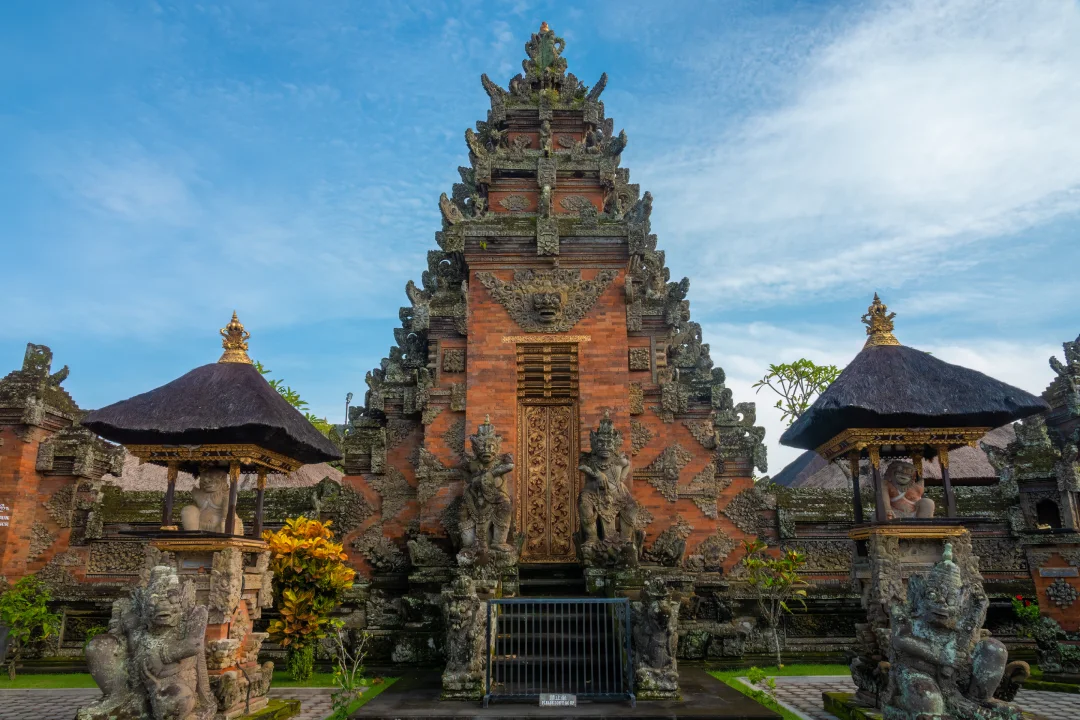 A traditional Balinese temple structure, with intricate carvings and statues. A prominent red-brick central gateway is adorned with ornate stonework, with guardian statues on either side, and all are surrounded by small shrines with thatched roofs under a bright, clear sky. 