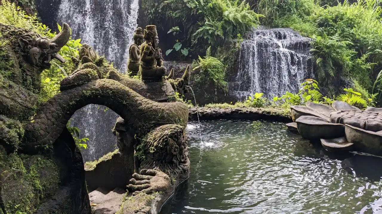 The foreground features intricately carved stone statues covered in moss, including a mythical dragon-like creature and meditative figures. In the background, greenery surrounds cascading waterfalls that fall into a pool.