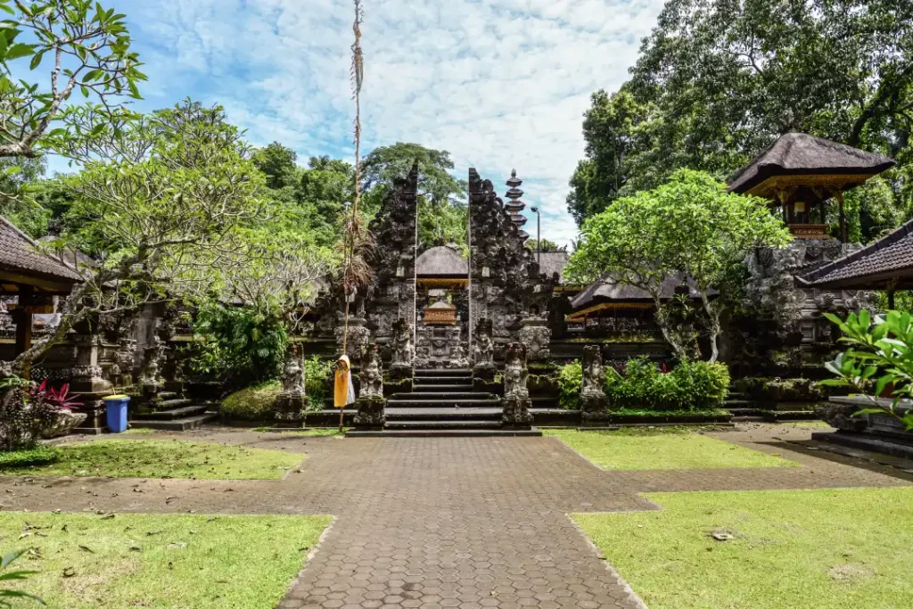 An ornate Balinese temple surrounded by greenery under a partly cloudy sky. The entrance features traditional stone carvings and intricate architecture, with small shrines and decorative elements typical of Balinese Hindu temples. 