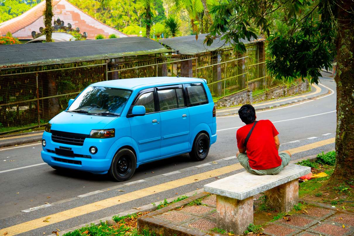 A bright blue minivan parked on a roadside in a tropical area in Bali. In the foreground, a man in a red shirt and khaki shorts is seen sitting on a concrete bench, facing away from the camera and observing the vehicle.