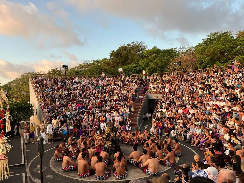 A large outdoor audience watching a traditional Balinese Kecak dance performance during sunset. In the centre are performers seated in a circular formation, chanting and gesturing, The setting is vibrant, with natural greenery and a clear, colourful sky in the background. 