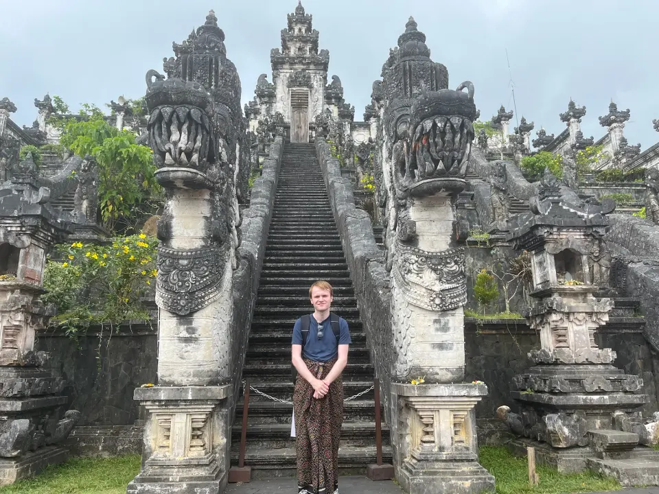 A person standing at the base of the staircase leading to a shrine at Lempuyang Temple in Bali, Indonesia. The ornate staircase has intricately carved stone dragons on either side and is surrounded by greenery and floral accents, with the temple structure visible at the top. 