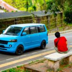 A bright blue minivan parked on a roadside in a tropical area in Bali. In the foreground, a man in a red shirt and khaki shorts is seen sitting on a concrete bench, facing away from the camera and observing the vehicle.