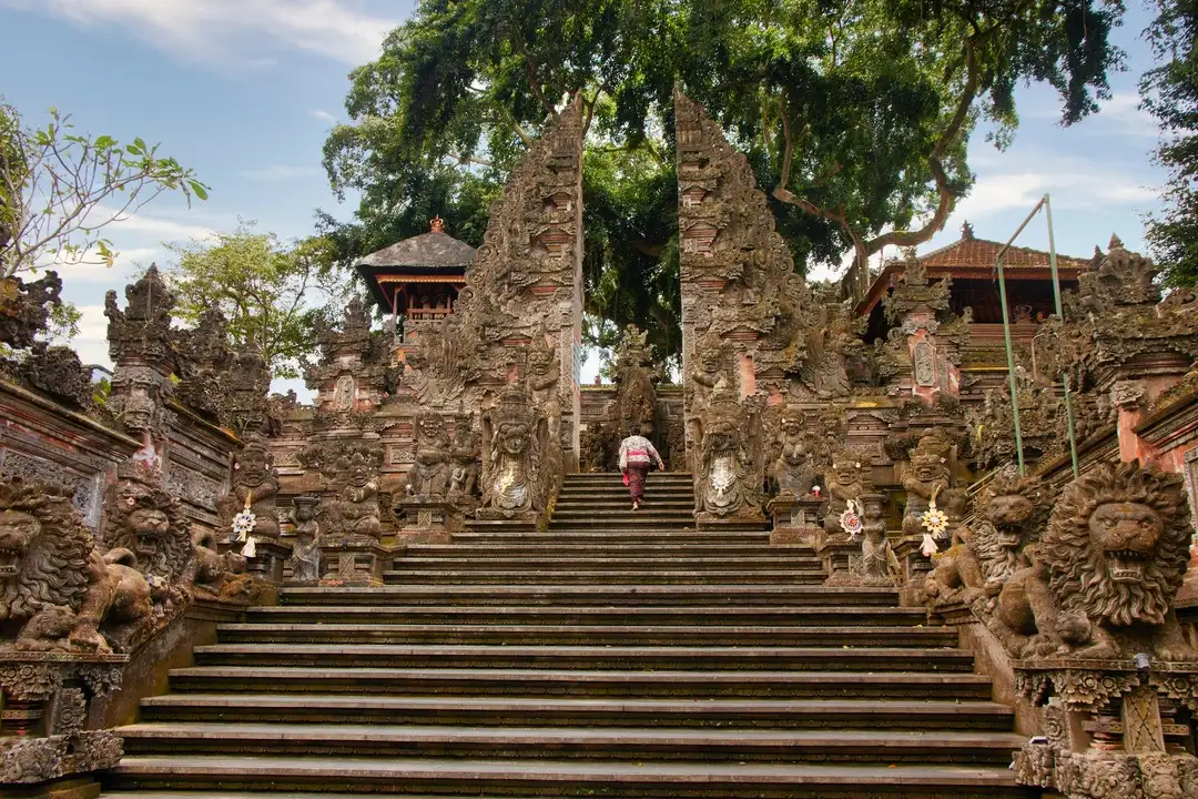 Entrance of Pura Dalem Ubud, a temple in Ubud, adorned with intricate stone carvings and statues. The stairway is flanked by guardian figures, with greenery and traditional pagoda-style roofs visible in the background