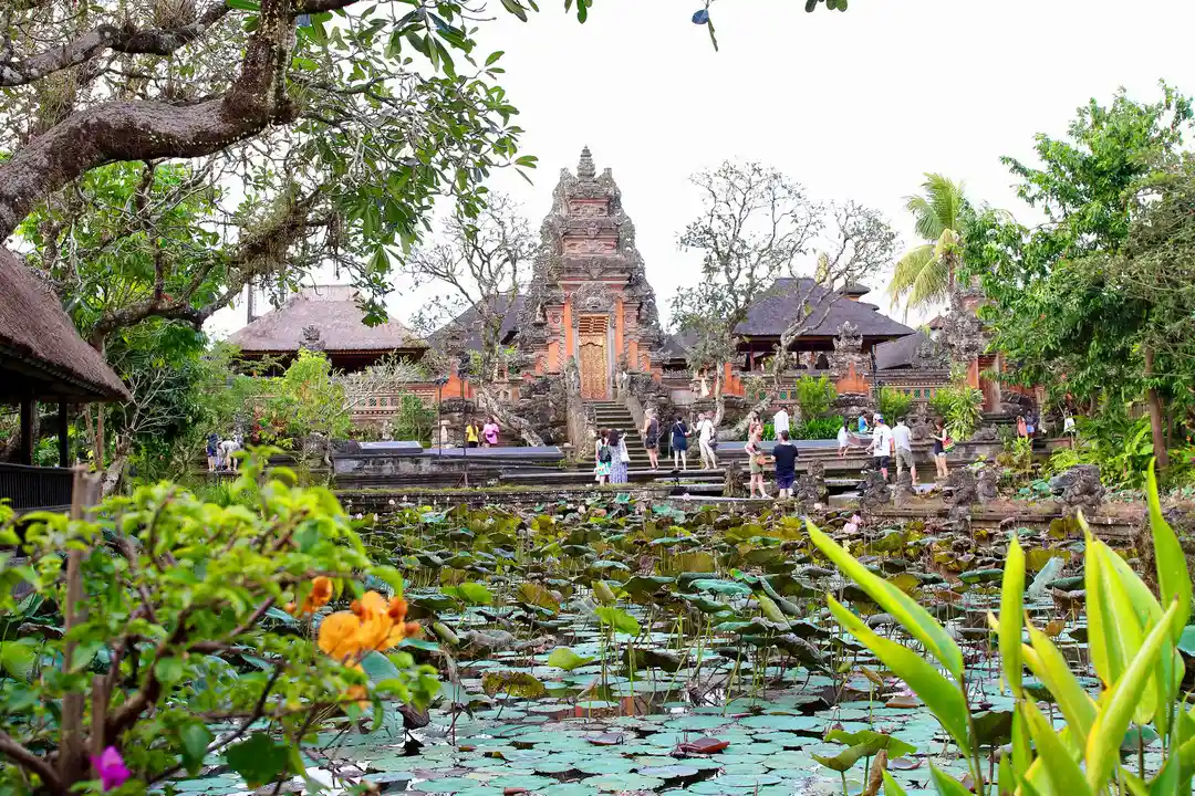 A tropical garden with a lotus pond in the foreground and Saraswati Temple in Ubud, Bali, in the background. Visitors are seen exploring the temple grounds, surrounded by ornate Balinese architecture and greenery.