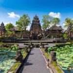 A view of the Saraswati Temple in Ubud, Bali. The temple is surrounded by greenery, lotus ponds, and Balinese architecture under a bright blue sky. The path leading to the temple is flanked by intricately carved stone statues.